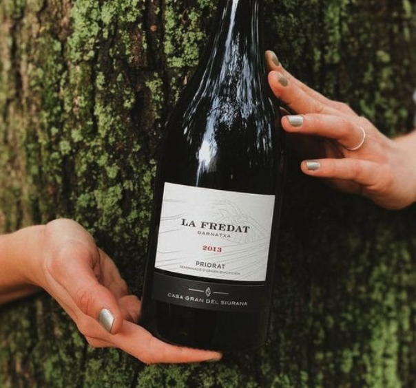 Two hands holding a bottle of La Fredat wine in a black bottle, against the bark of a treetrunk.